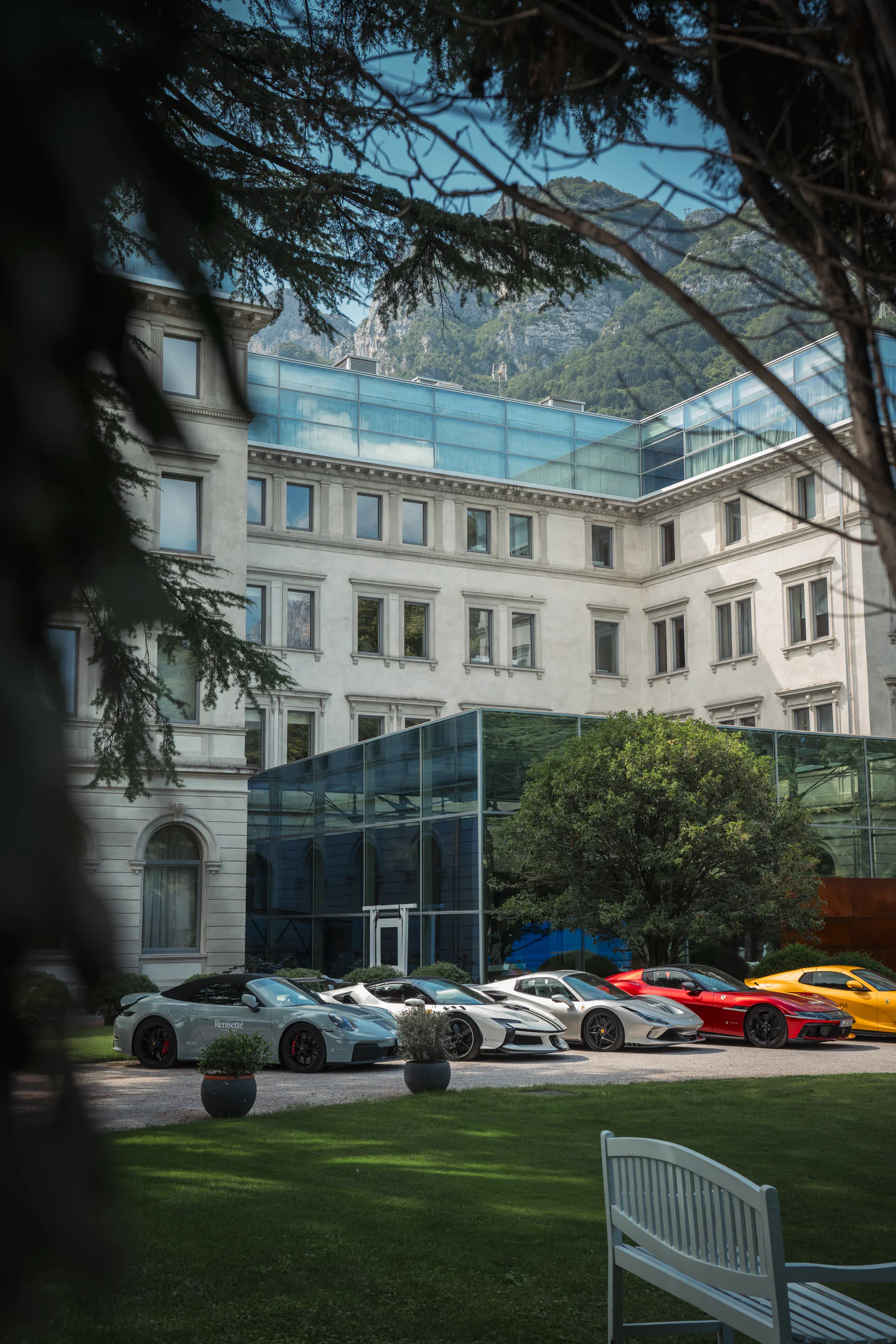 Porsche 911s, a red Ferrari and a yellow Ferrari parked outside a grand hotel beneath the mountains