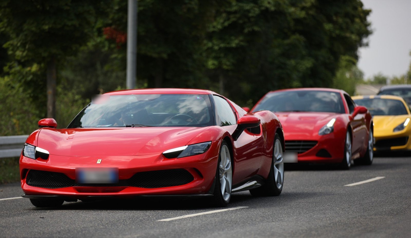 Red Ferrari 296 GTB leading a convoy of Ferraris along a tree-lined road
